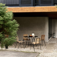 "Black metal frame dining set with light wood slats, set against a neutral stone wall and surrounded by greenery."