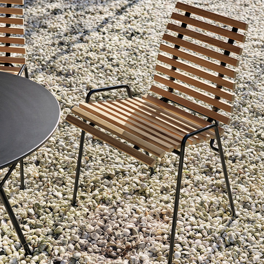 Wooden slatted chair with a natural finish, paired with a sleek black round table against a pebbled outdoor setting.