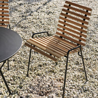 Wooden slatted chair with a natural finish, paired with a sleek black round table against a pebbled outdoor setting.