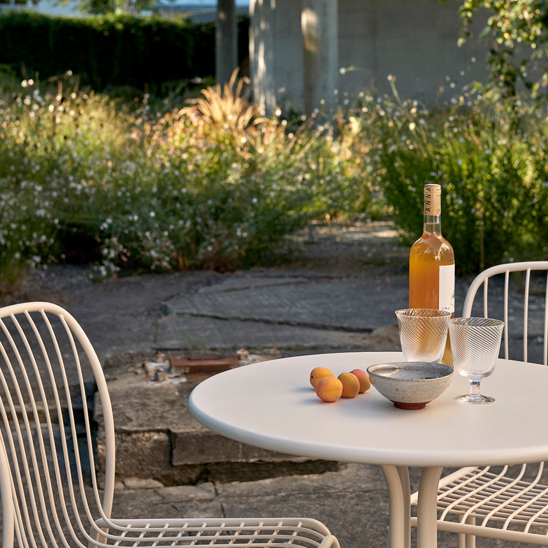 "White round table set with clear glasses and a beige bottle, surrounded by lush green plants and subtle floral patterns."
