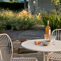 "White round table set with clear glasses and a beige bottle, surrounded by lush green plants and subtle floral patterns."