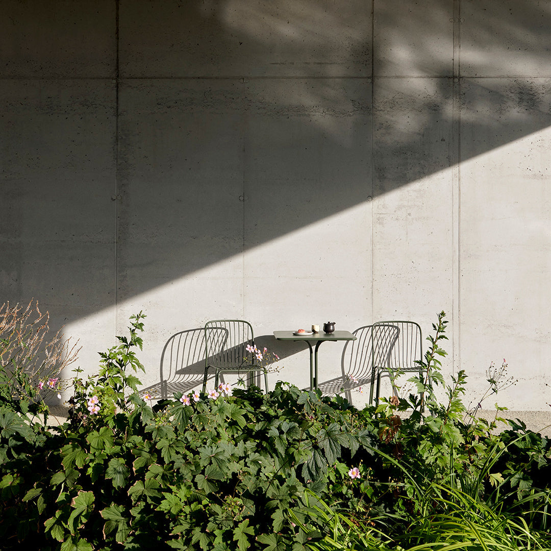 Shadowed concrete wall with green foliage and pink flowers, showcasing modern black wire chairs and a minimalist table.