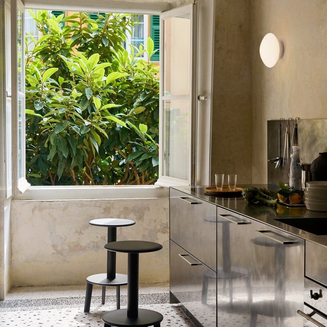 Black round stools contrasting with a sleek metallic kitchen, framed by light greenery outside a bright window.