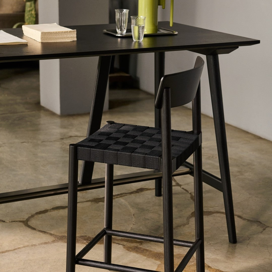 Black woven chair with a sleek design against a minimalist gray background and subtle stone floor patterns.