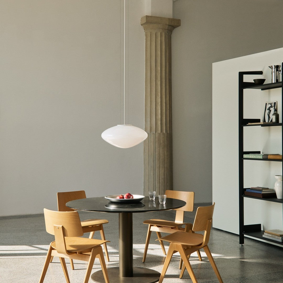Sleek white pendant light above a circular black table, surrounded by light wooden chairs in a minimalist space.