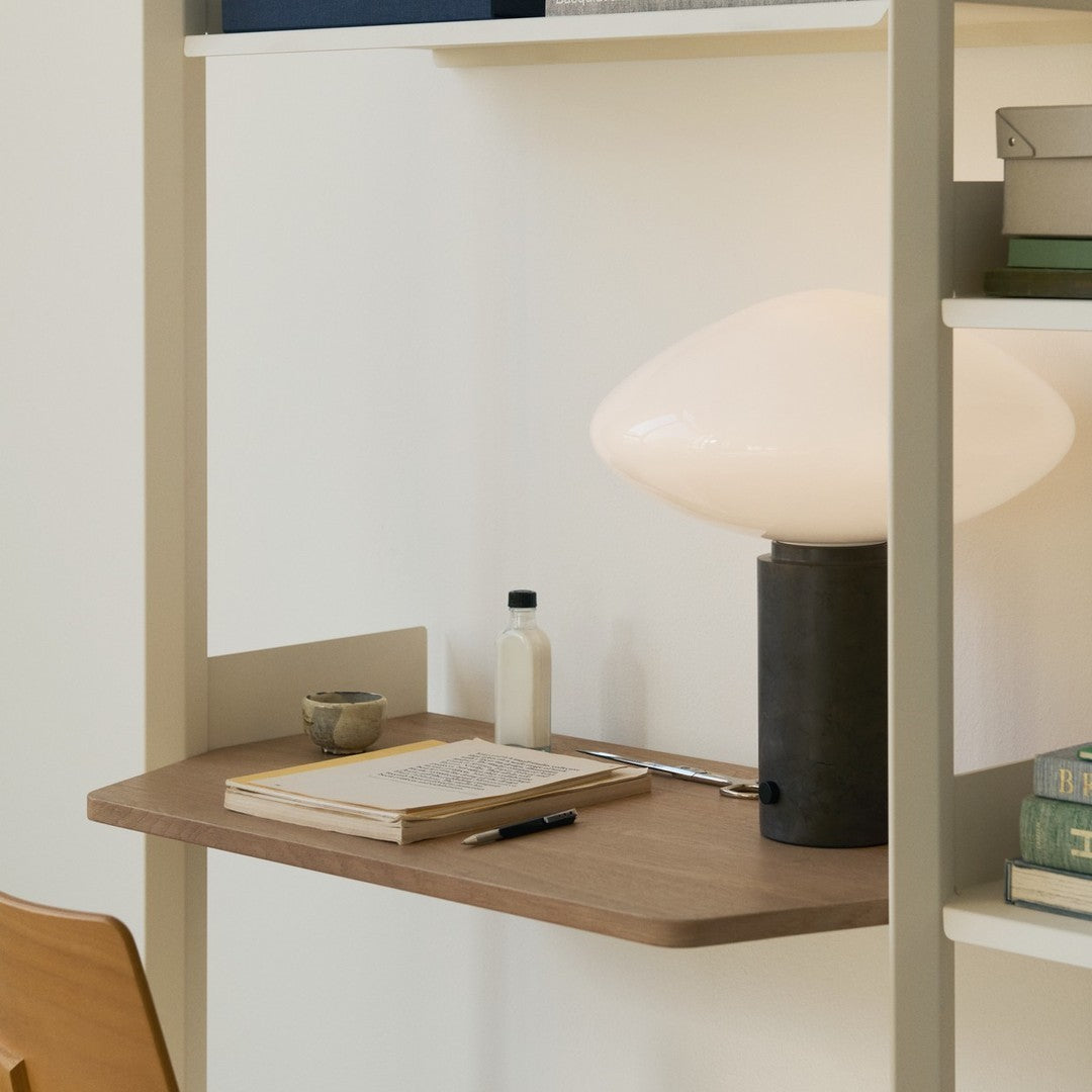 Wooden desk with light beige shelves, black lamp, white glass shade, and blue-green books create a stylish, modern workspace.