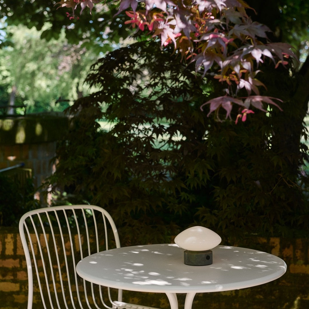 White metal chair and table with a smooth, oval lamp against green foliage and vibrant red-purple leaves.