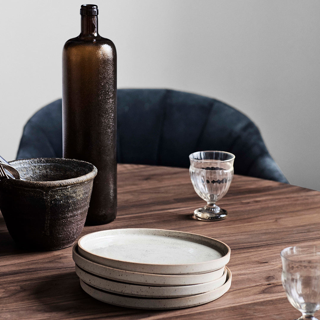 Brown glass bottle, textured ceramic bowl, clear glassware, and white plates on a wooden table with a dark blue chair.