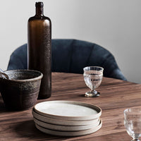 Brown glass bottle, textured ceramic bowl, clear glassware, and white plates on a wooden table with a dark blue chair.