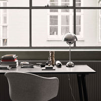 "Modern workspace with gray chair, black desk, silver lamp, and stacked books, featuring neutral tones and sleek lines."