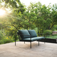 Teal cushioned outdoor sofa with black metal frame, set against a lush green backdrop and warm sunlight.