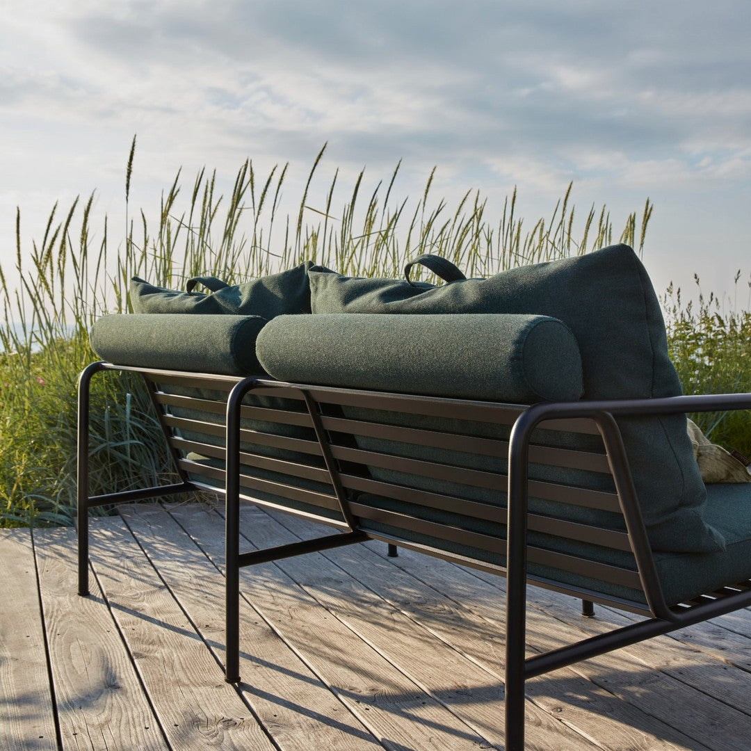 Green cushion sofa with black metal frame, set against a natural backdrop of tall grass and a cloudy sky.