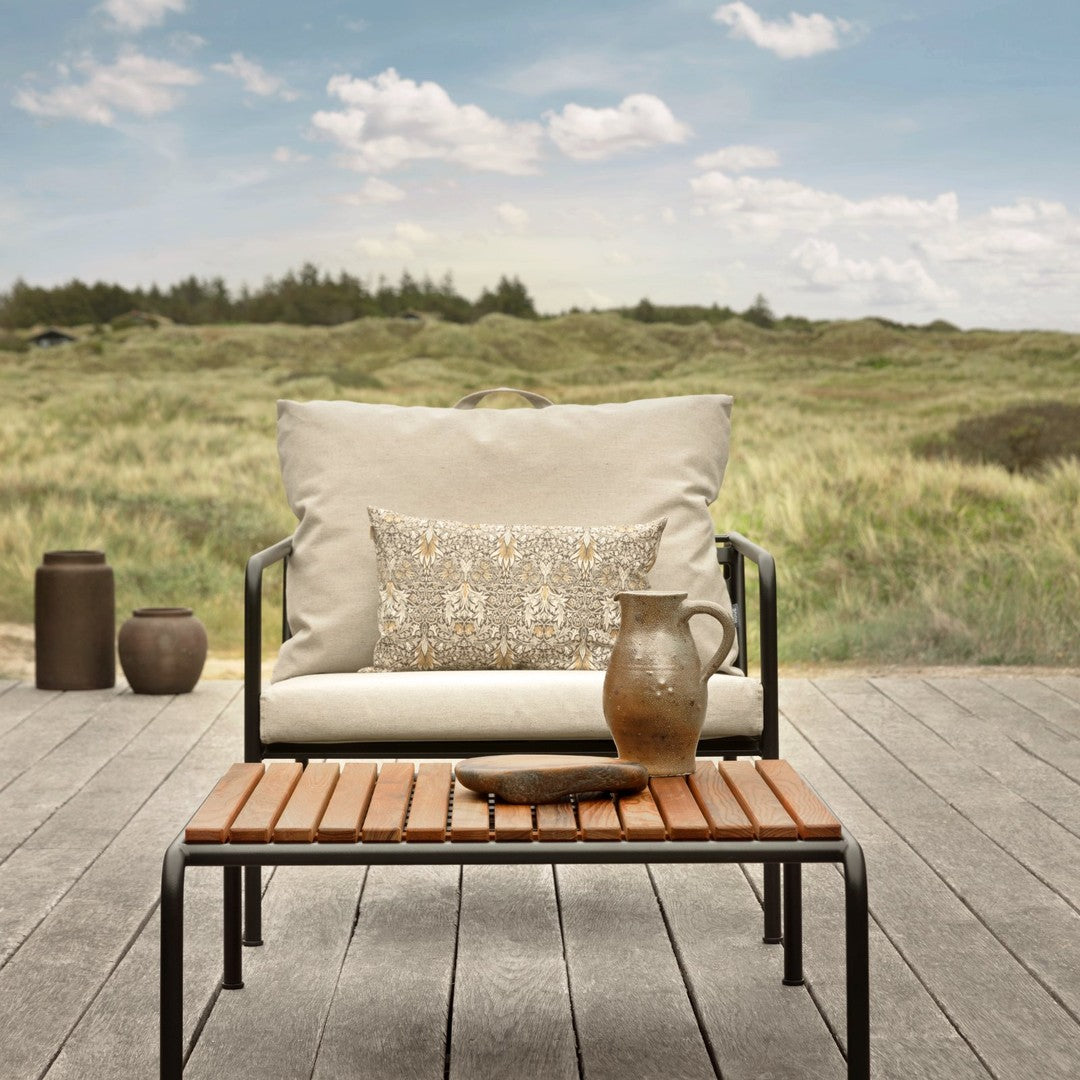 Minimalist outdoor lounge chair with cream cushions and decorative earthy-toned pillow, set against a lush green landscape.