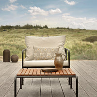 Minimalist outdoor lounge chair with cream cushions and decorative earthy-toned pillow, set against a lush green landscape.