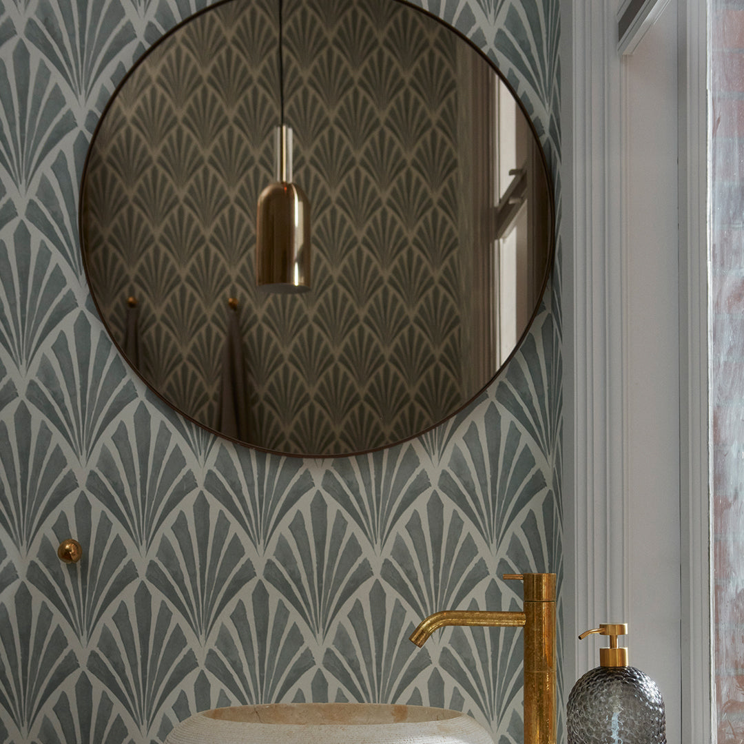 Round mirror with a gold frame against a teal wallpaper featuring a stylish art deco pattern in muted tones.