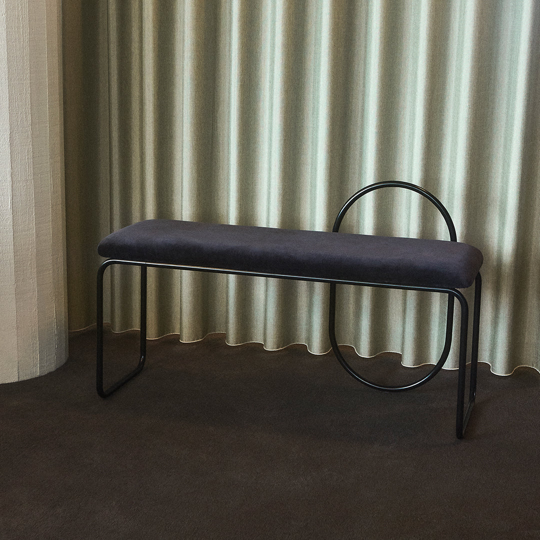Gray upholstered bench with black metal legs and unique circular accent against soft beige curtains.