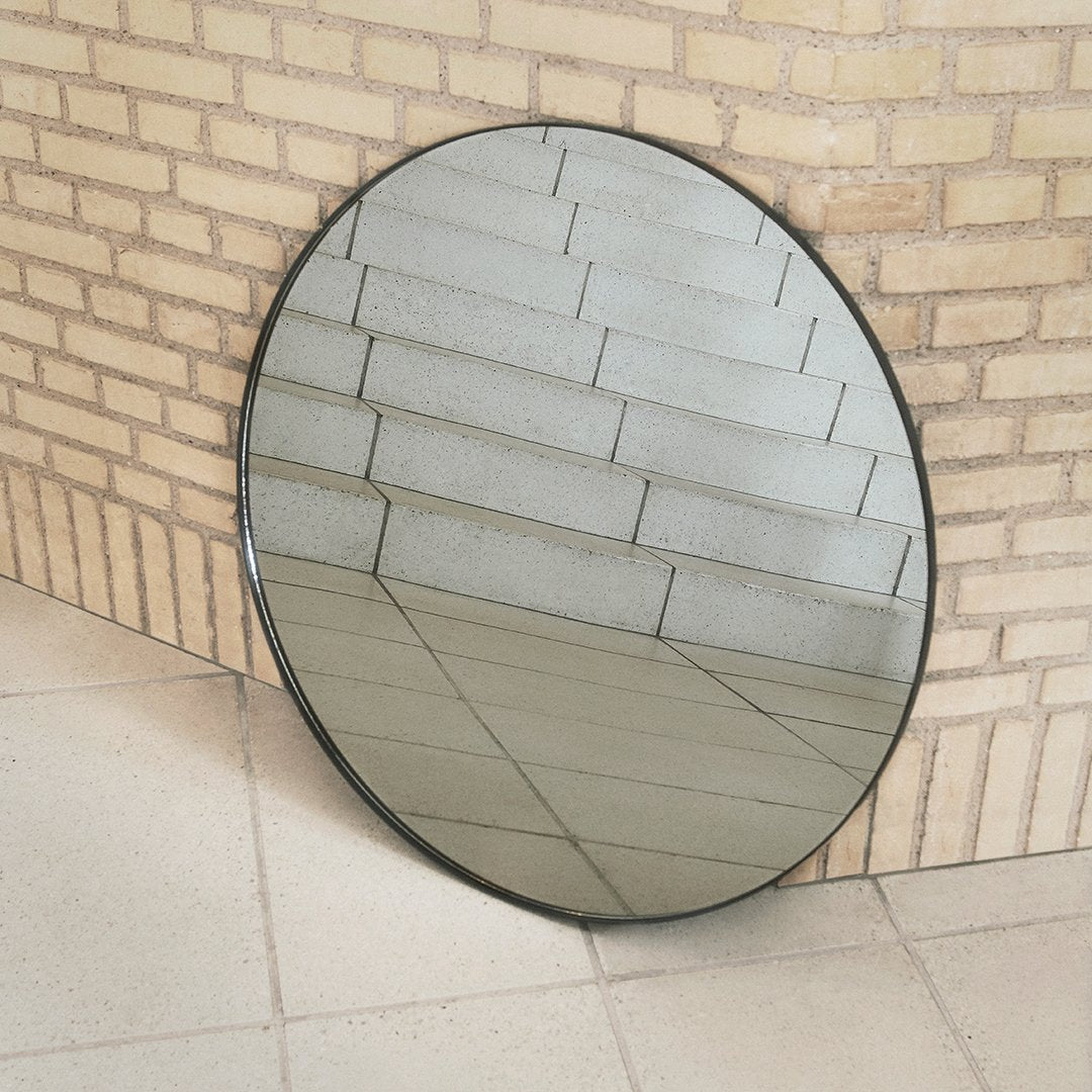 Round black-framed mirror reflecting textured gray brick wall and floor tiles, showcasing sleek design and modern aesthetics.