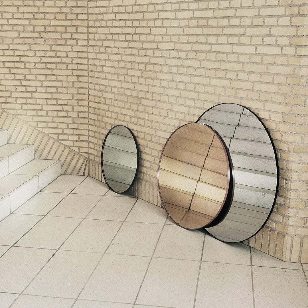 Modern circular mirrors in silver, bronze, and transparent finishes against a textured beige brick wall.