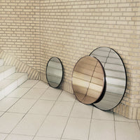 Modern circular mirrors in silver, bronze, and transparent finishes against a textured beige brick wall.