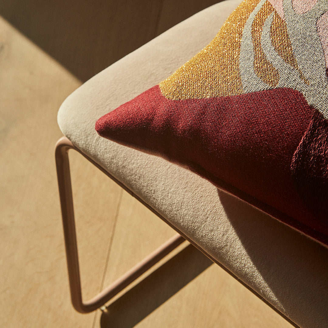 Decorative pillow with abstract patterns in burgundy, gold, and cream on a soft beige cushion. Perfect for modern décor.