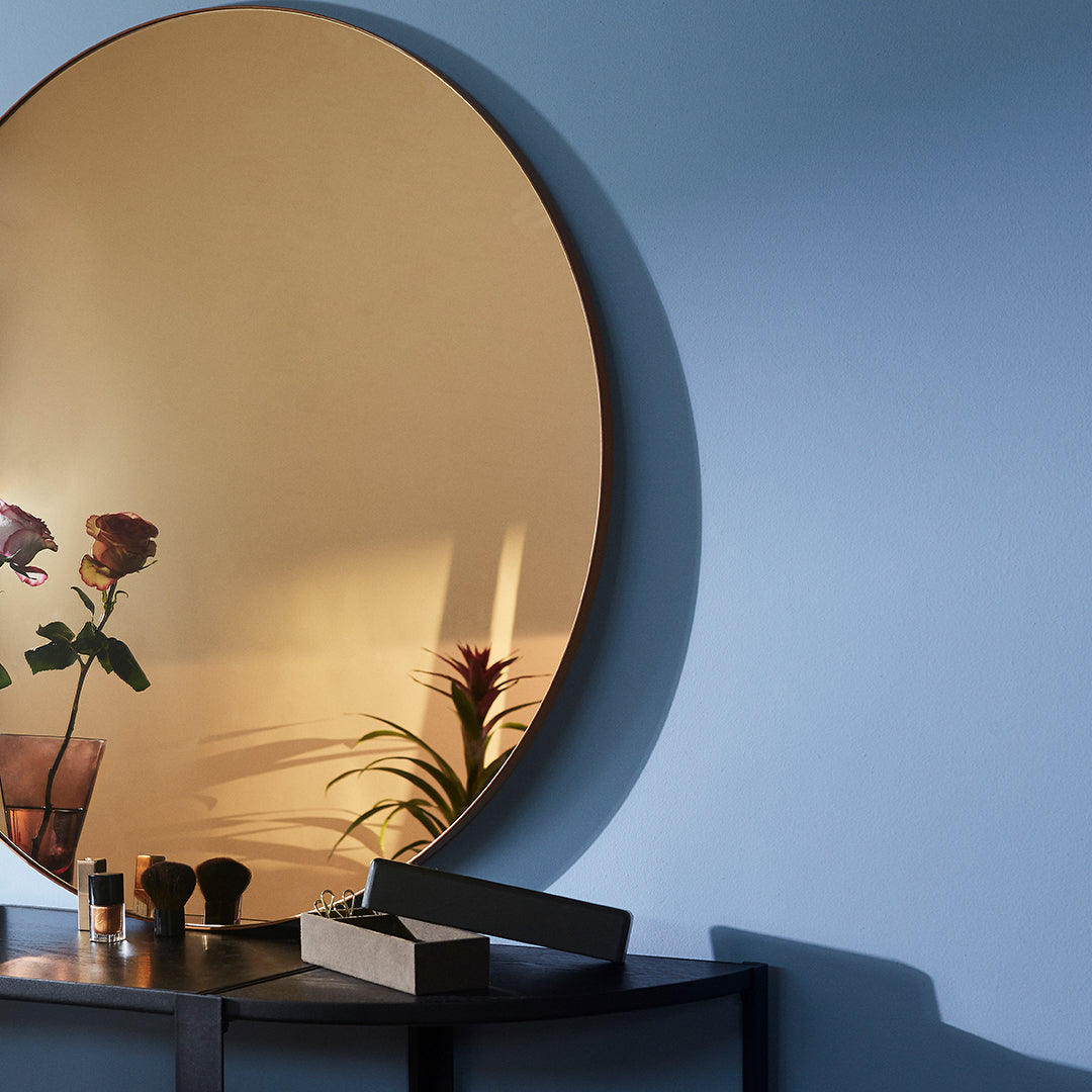 Round gold mirror reflecting soft blue wall, with hints of green from plants and pink roses, creating a serene ambiance.