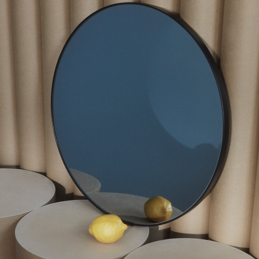 Round black-framed mirror with a reflective dark blue surface, set against neutral cylindrical backdrops.