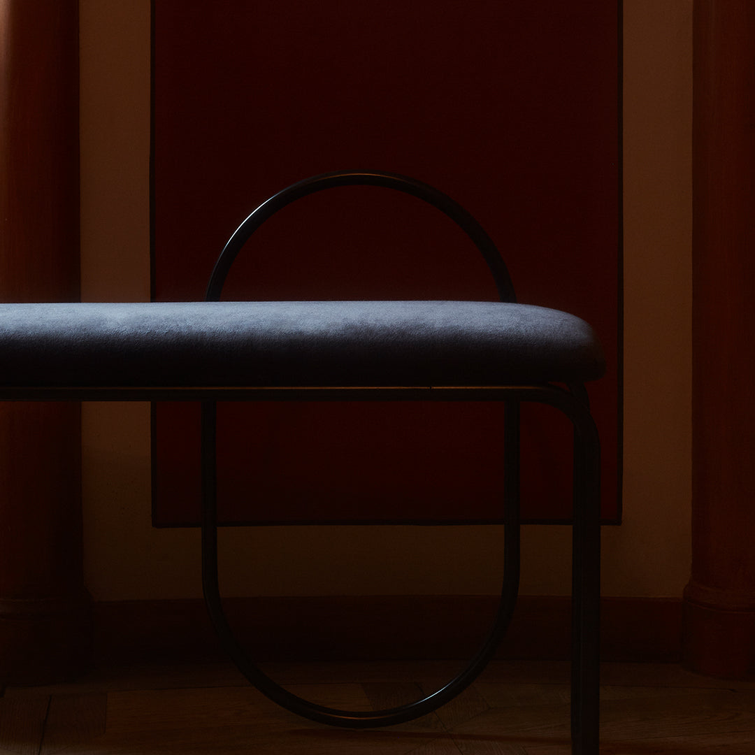 Gray upholstered bench with sleek black metal frame, set against a deep red wall. Minimalist, modern design.