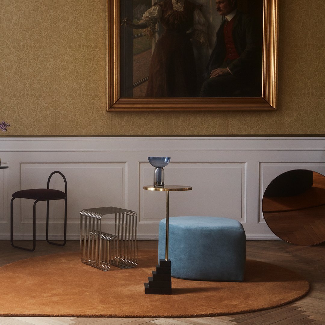 Soft blue ottoman, clear side table, and dark brown rug in an elegant room with vintage wallpaper and artwork.