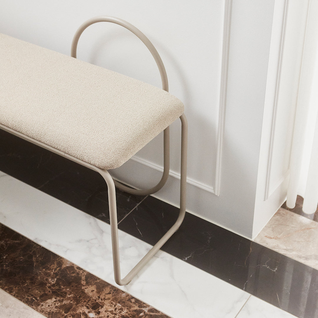 Beige upholstered bench with curved metal frame, set on a contrasting marble floor featuring black and cream patterns.