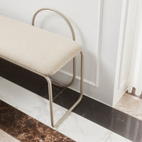 Beige upholstered bench with curved metal frame, set on a contrasting marble floor featuring black and cream patterns.
