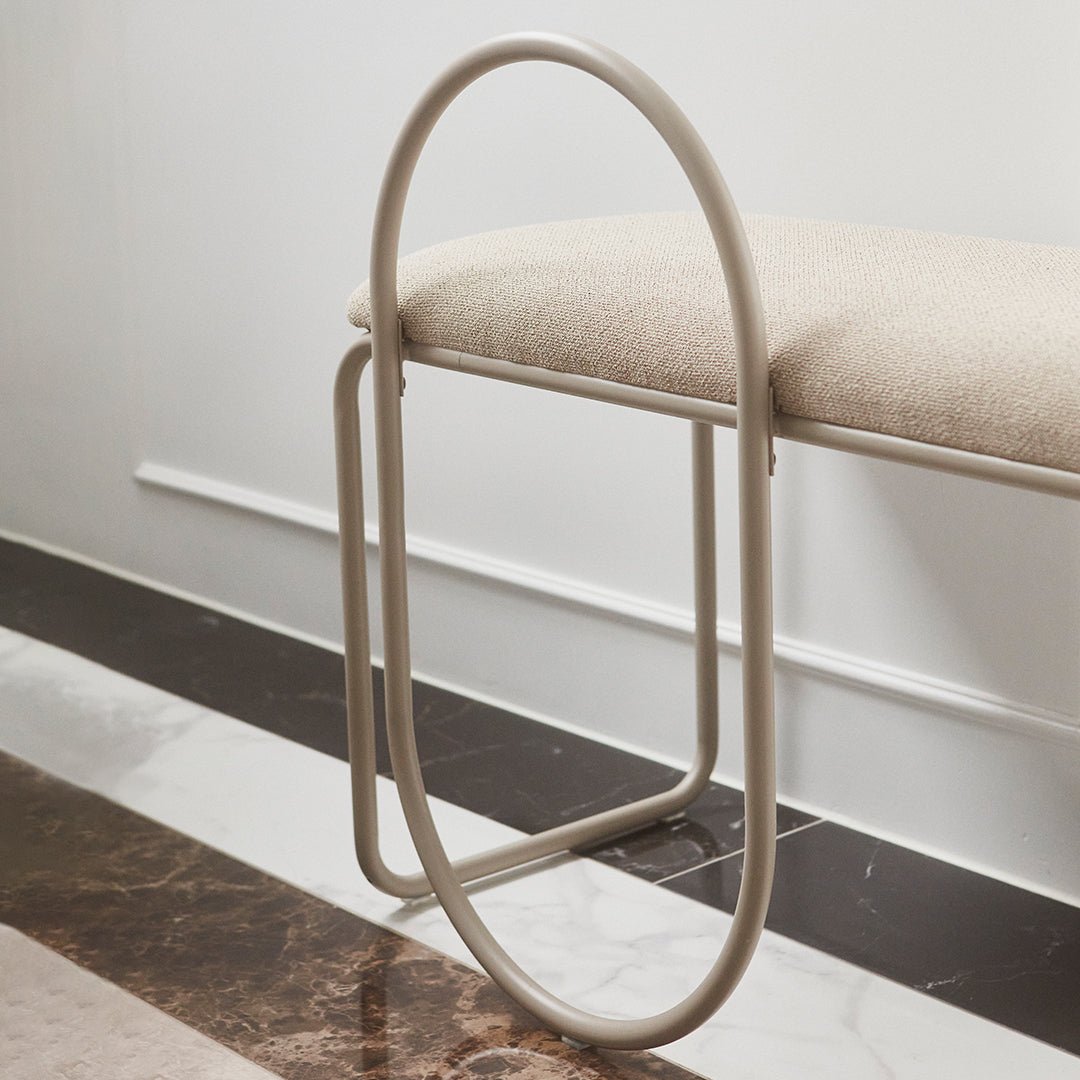 Beige upholstered bench with a minimalist oval metal frame, set against a sleek black and white marble floor.