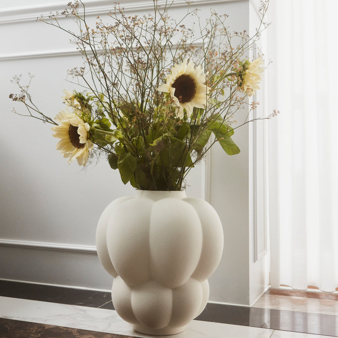 White textured vase with circular patterns, filled with sunflowers and greenery, set against a light, airy background.