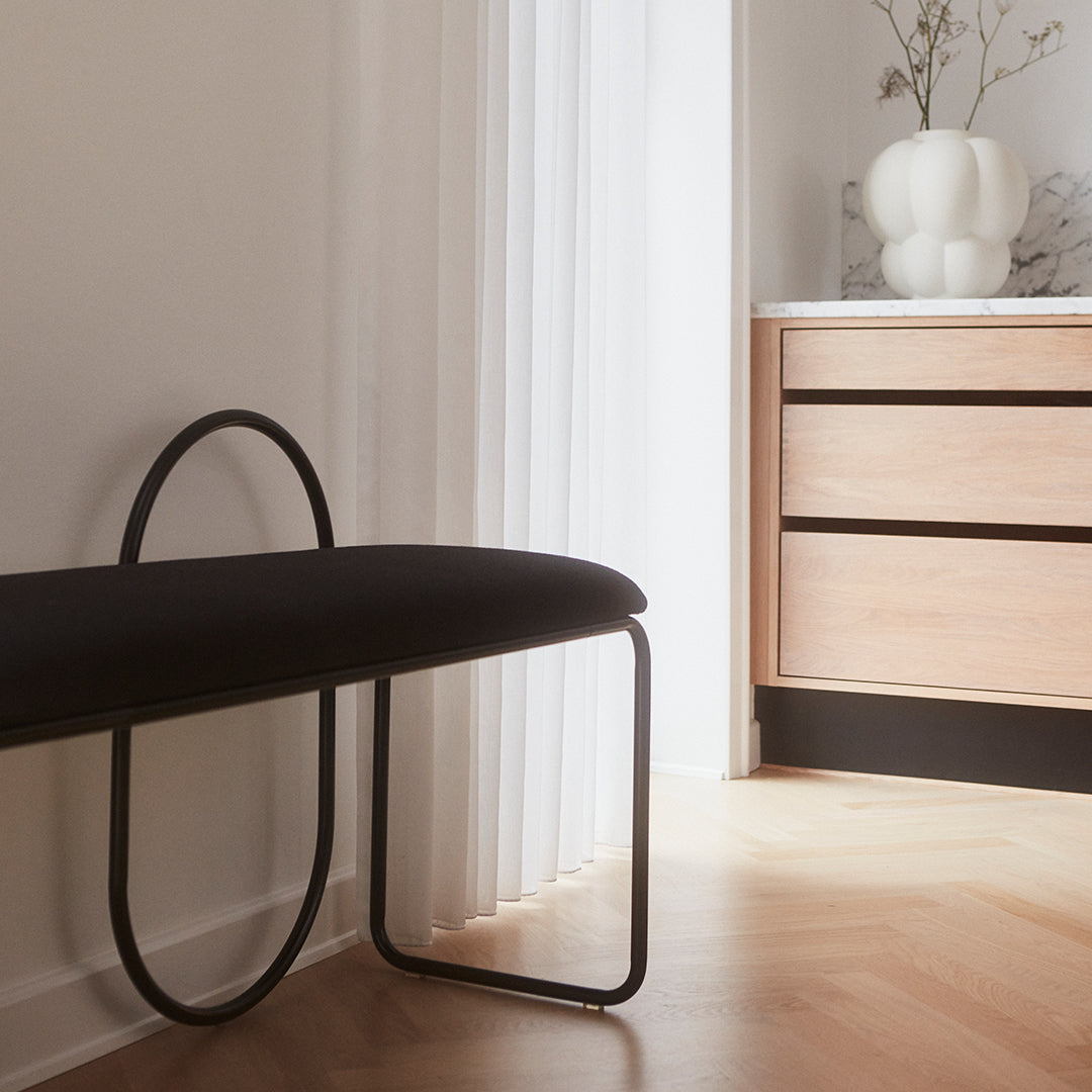 Black upholstered bench with a sleek curved metal frame on light wood flooring, complemented by neutral decor.