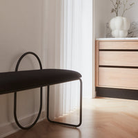 Black upholstered bench with a sleek curved metal frame on light wood flooring, complemented by neutral decor.