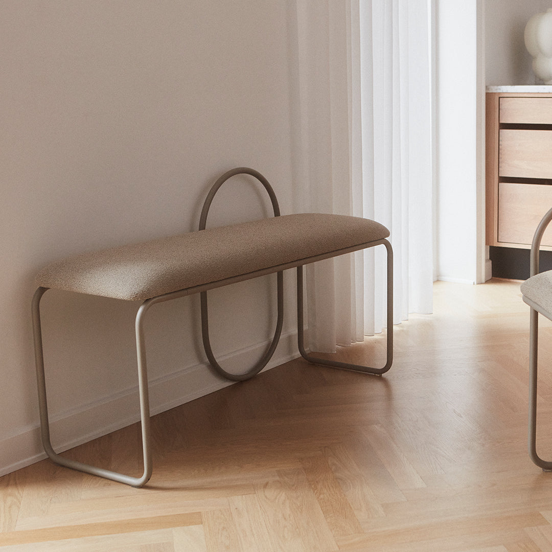 Beige upholstered bench with a minimalist silver frame, featuring smooth curves and a contemporary design.