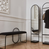 Black bench and mirror with sleek metal frames, set against a light-colored wall and wooden herringbone floor.