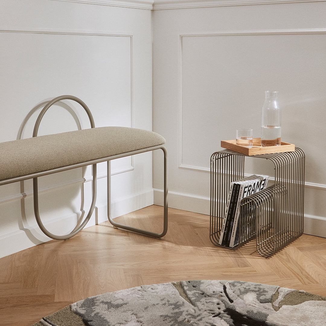 "Beige upholstered bench with curved metal legs, paired with a sleek wire side table and a patterned round rug."