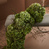 Lush green floral arrangement with textured layers, accented by delicate branches against a soft pink background.