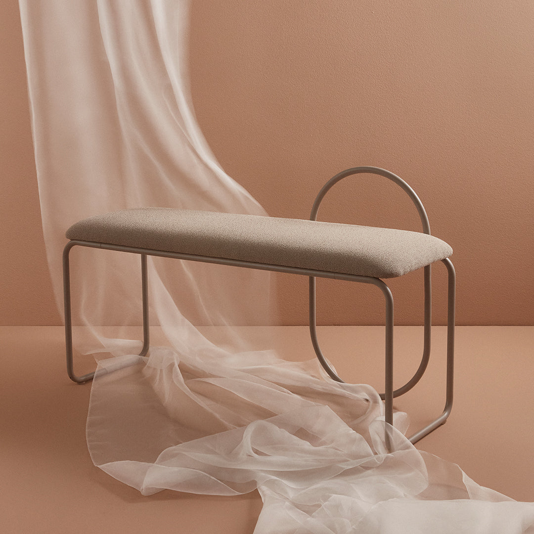 Light beige upholstered bench against a warm, muted peach backdrop, draped with soft, sheer white fabric.