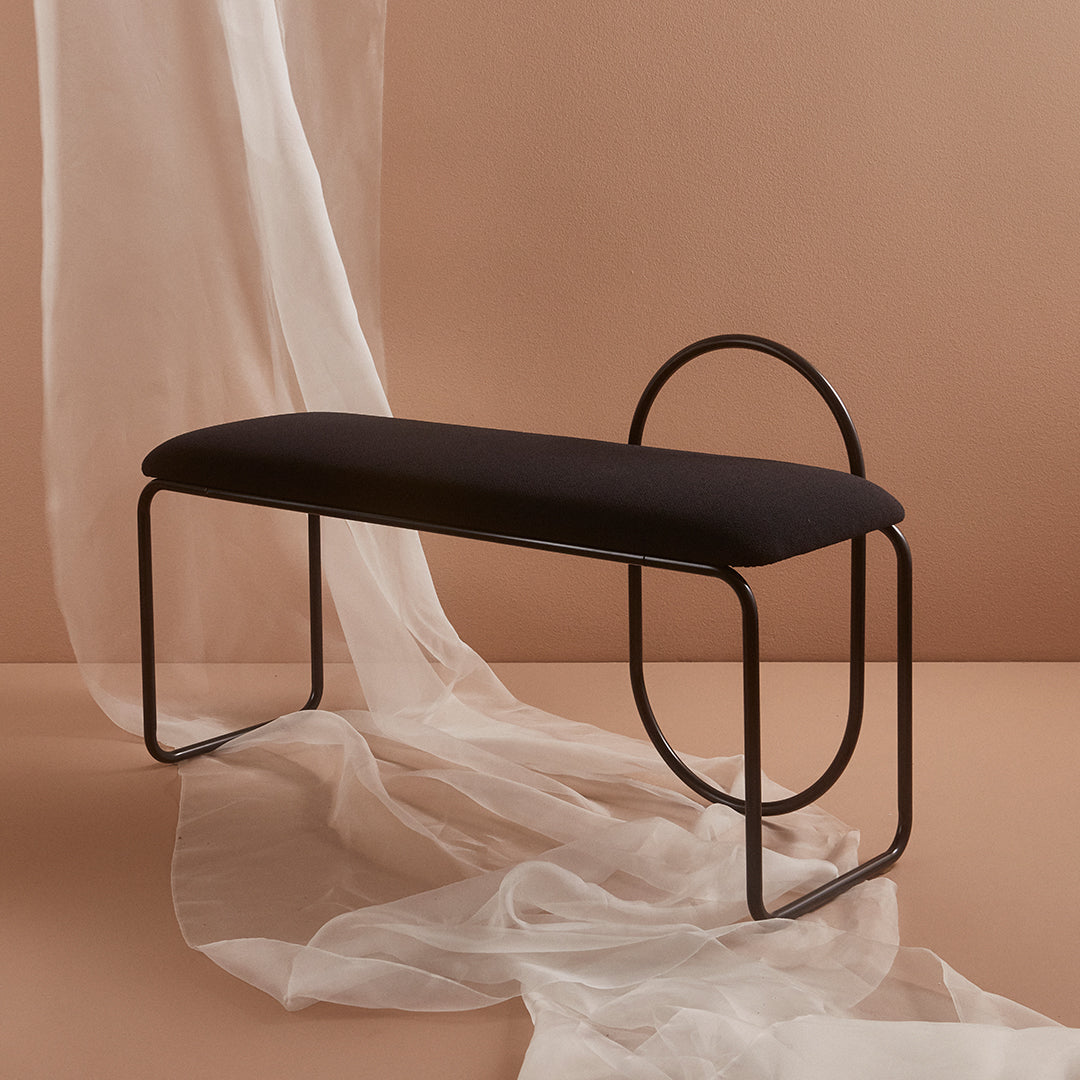 Black upholstered bench with minimalist metal frame on a soft beige background and flowing sheer fabric.