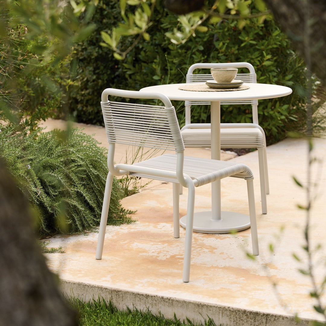 Stylish light gray outdoor dining set with textured chair and smooth round table, set against lush greenery.