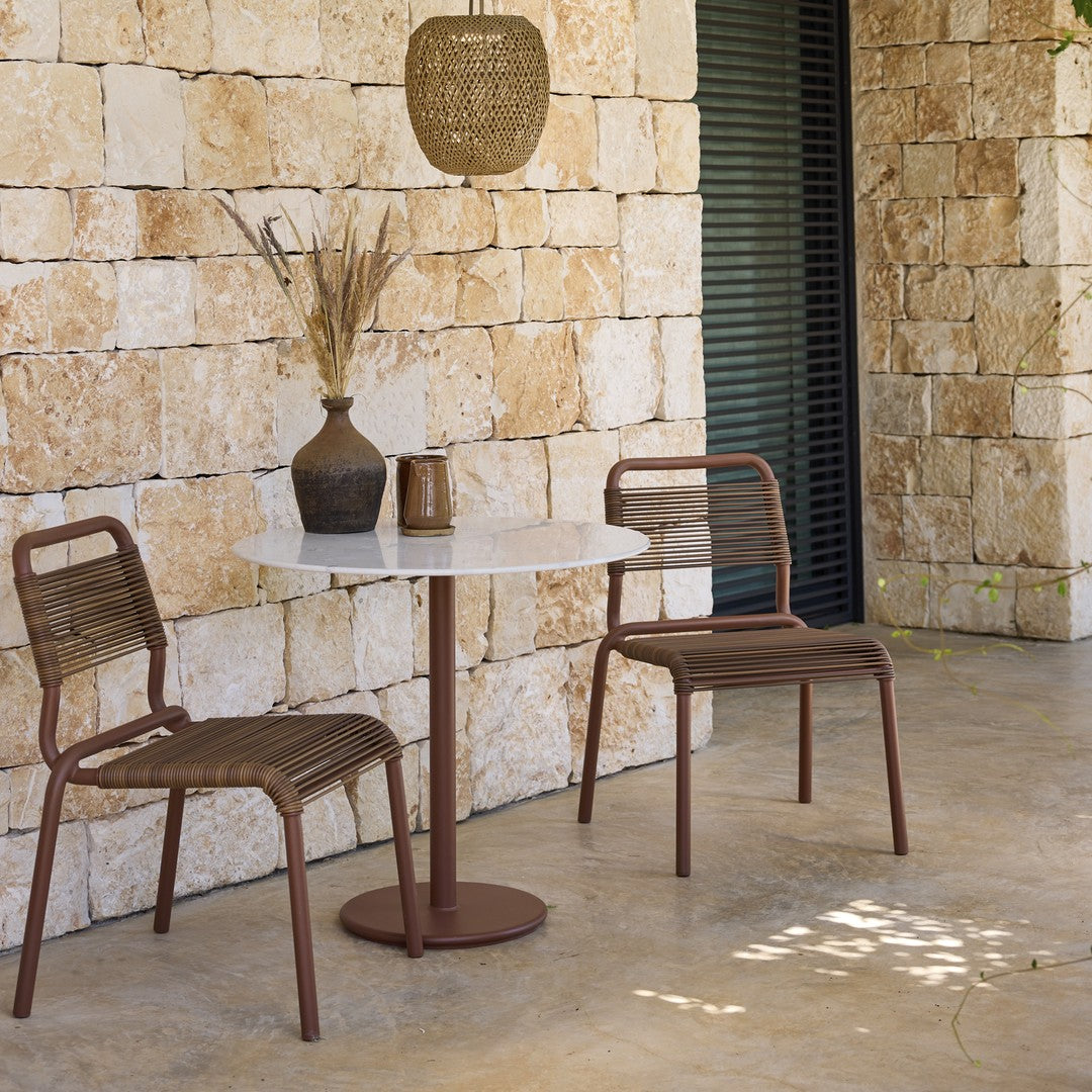 Brown chairs with woven patterns and a white marble table against a textured stone wall, creating a modern outdoor setting.