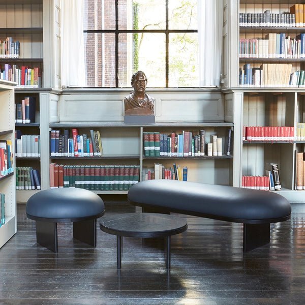 Black curved bench and table set on dark hardwood floor, surrounded by a library with colorful bookshelves.