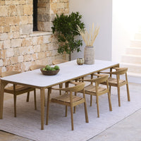 Light wood dining table with natural finish, paired with beige chairs on a textured neutral rug against a stone wall.