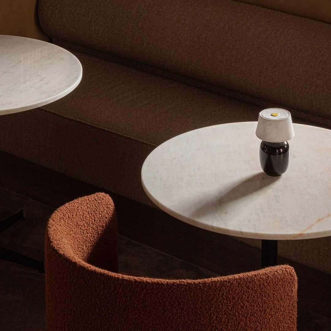 Textured rust-colored chair beside round marble tables in a warm-toned, minimalist setting. Soft natural light.