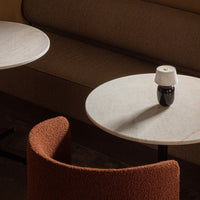Textured rust-colored chair beside round marble tables in a warm-toned, minimalist setting. Soft natural light.