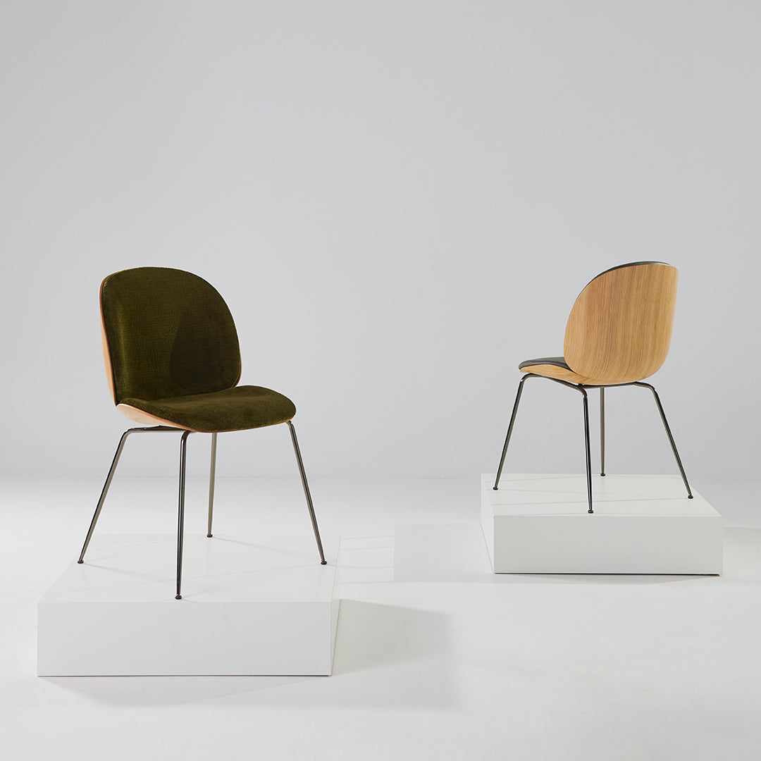 Olive green velvet chair with natural wood back and black metal legs, displayed on a white pedestal. Minimalist design.