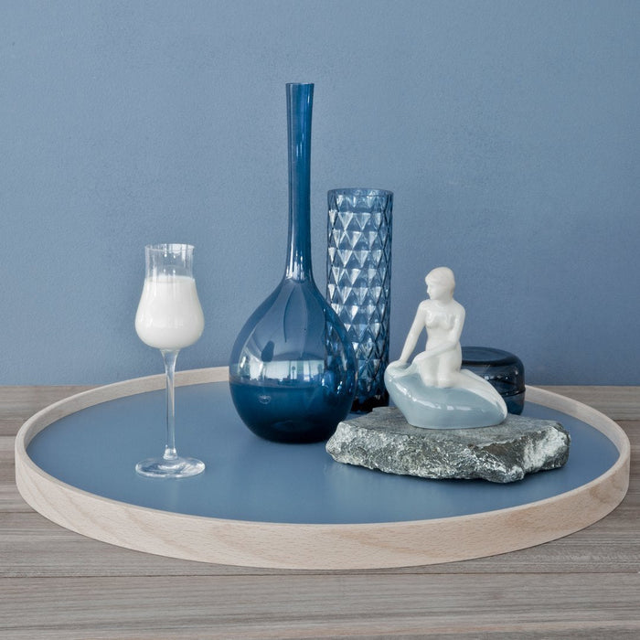 Blue glass vases, a white figurine, and a textured stone on a round wood tray, set against a soft blue wall.
