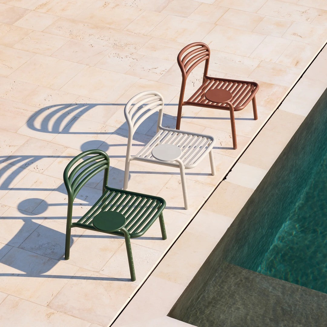 "Three modern chairs in green, white, and rust with a slatted design, placed by a pool on a stone patio."
