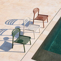 "Three modern chairs in green, white, and rust with a slatted design, placed by a pool on a stone patio."